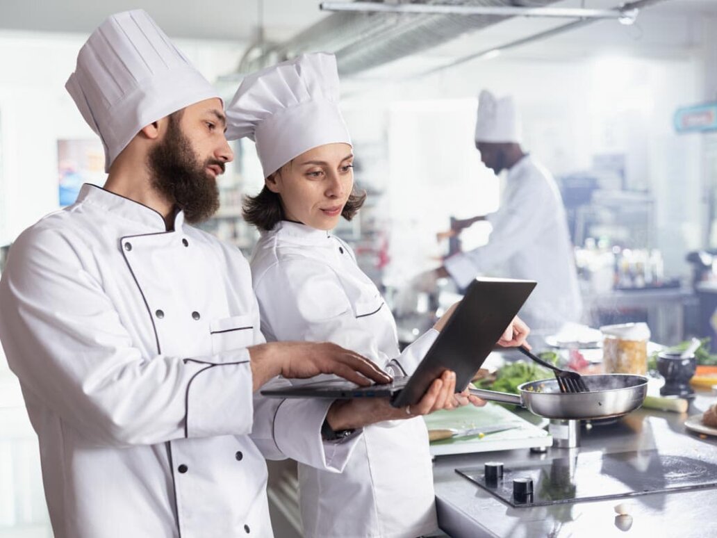 Zwei Köche planen die Warenwirtschaft mit dem Necta Group System. Sie optimieren Bestellprozesse in einer großen Edelstahl-Profi-Restaurantküche.