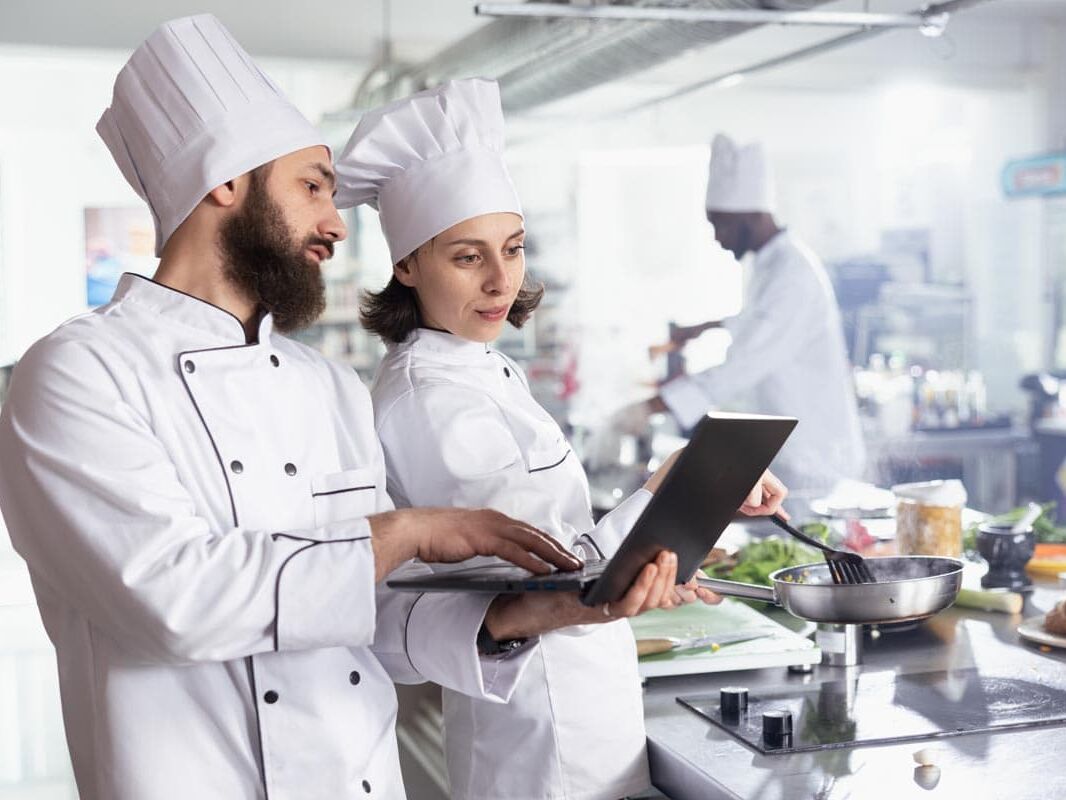 Zwei Köche planen die Warenwirtschaft mit dem Necta Group System. Sie optimieren Bestellprozesse in einer großen Edelstahl-Profi-Restaurantküche.