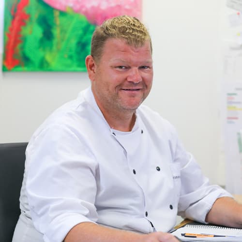 Mathias Rueßler, IN VIA Köln, Portraitfoto in weißer Kochuniform am Schreibtisch